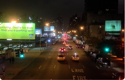 Night-time city road with multiple vehicles, illuminated streetlights, and digital advertising signs along the roadside.